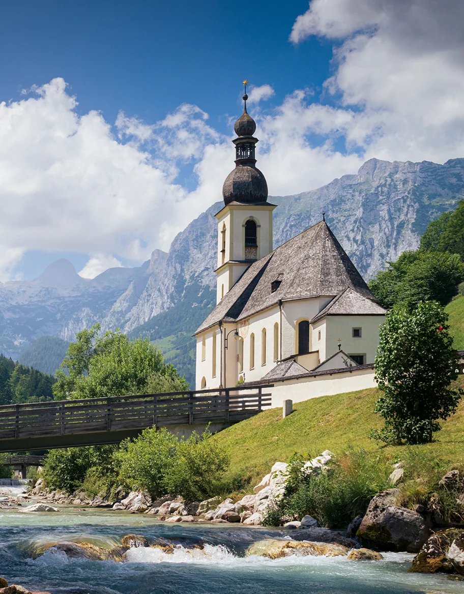 Eine Kirche an einem Gebirgsbach, dahinter Berge und Wolken