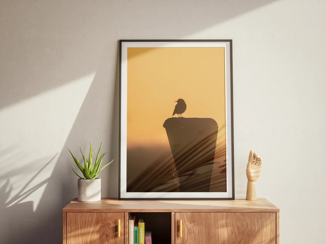 Der gerahmte Druck mit einer Vogelsilhouette vor orangem Abendhimmel steht auf einem Sideboard neben einer Pflanze