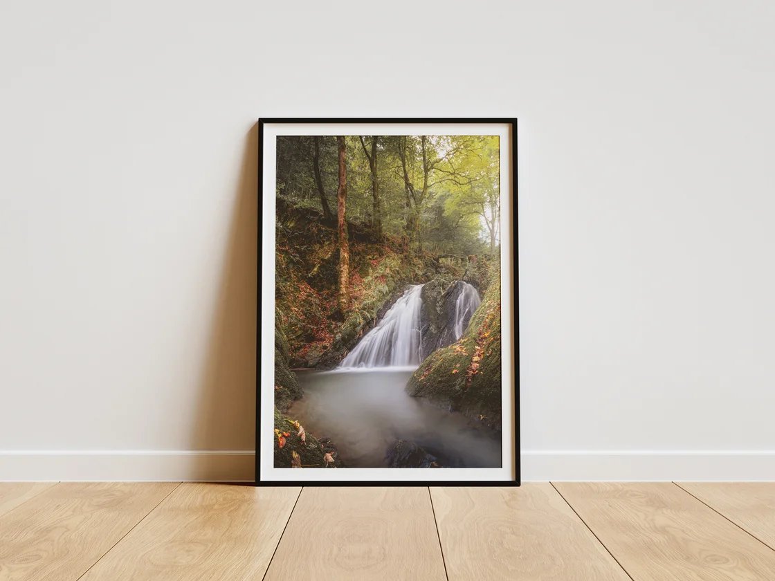 Der gerahmte Druck eines Wasserfalls im herbstlichen Wald lehnt an einer hellen Wand auf hellem Holzboden