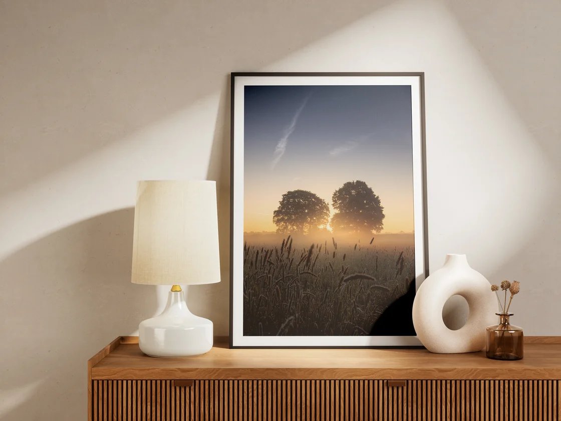 Der gerahmte Druck mit zwei Eichen im Morgenlicht über einem Feld steht auf einem Sideboard neben einer Lampe