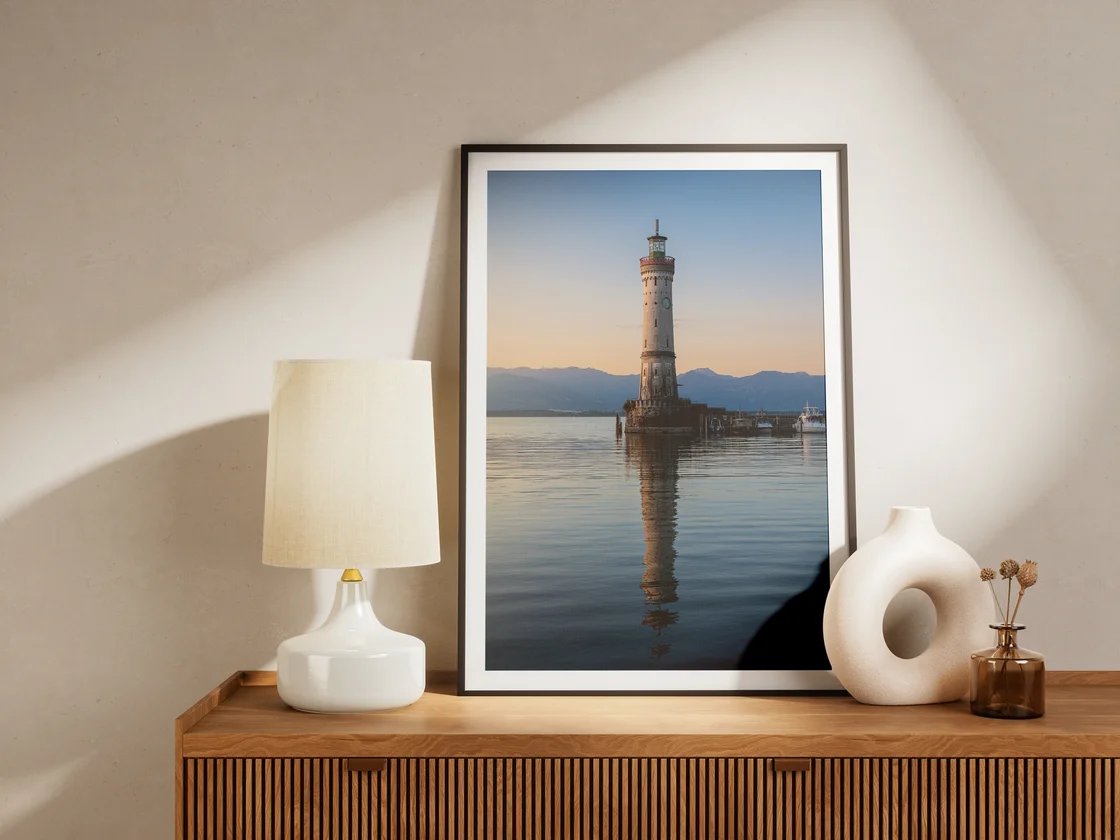 Der gerahmte Druck des Lindauer Leuchtturms, der sich im ruhigen Wasser spiegelt, steht auf einem Sideboard zwischen Lampe und Vase