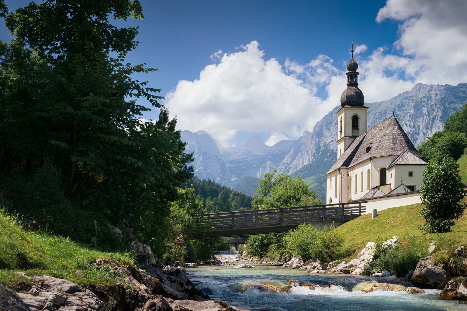 Eine Kirche an einem Gebirgsbach, dahinter Berge und Wolken