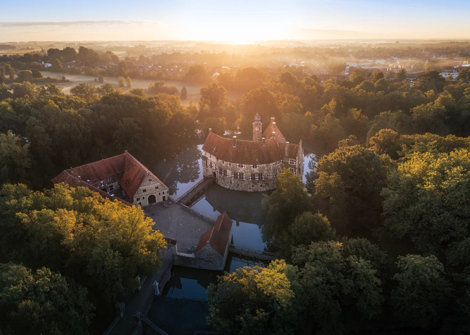 Luftaufnahme einer Wasserburg zwischen Bäumen, dahinter geht die Sonne auf