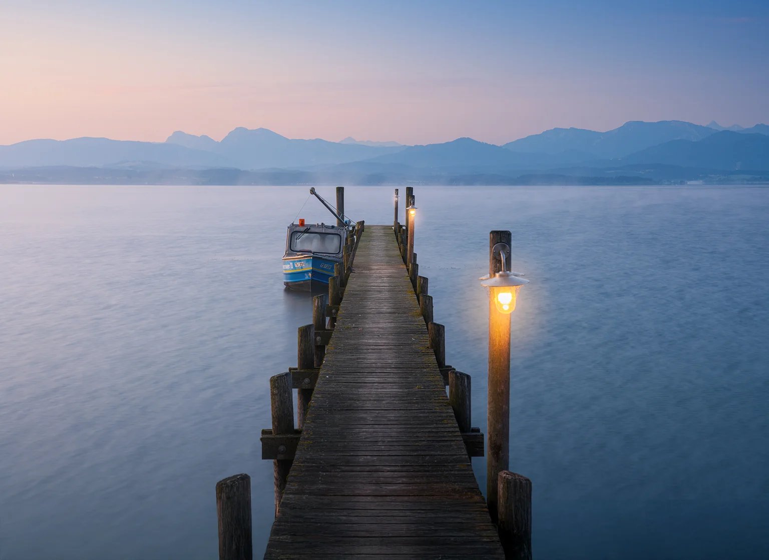 Ein Holzsteg mit Laterne führt in einen ruhigen See, dahinter Berge in der Morgendämmerung