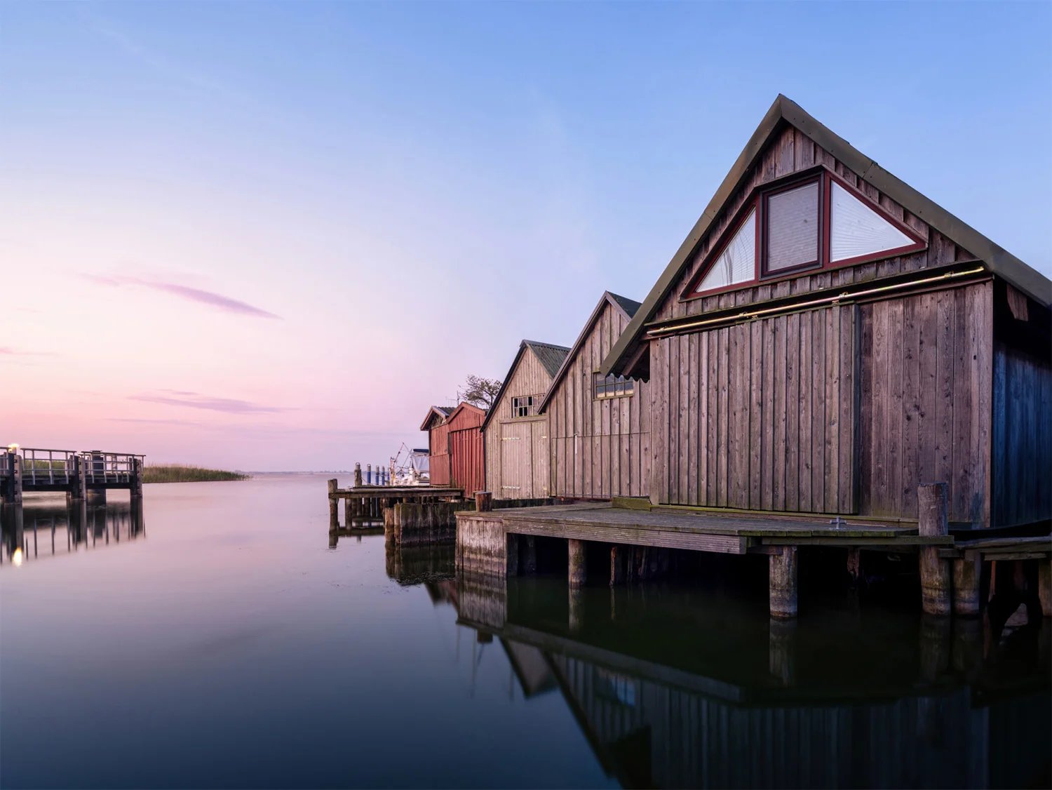 Hölzerne Bootshäuser stehen auf Stegen im stillen Wasser, darüber ein rosa-blauer Abendhimmel