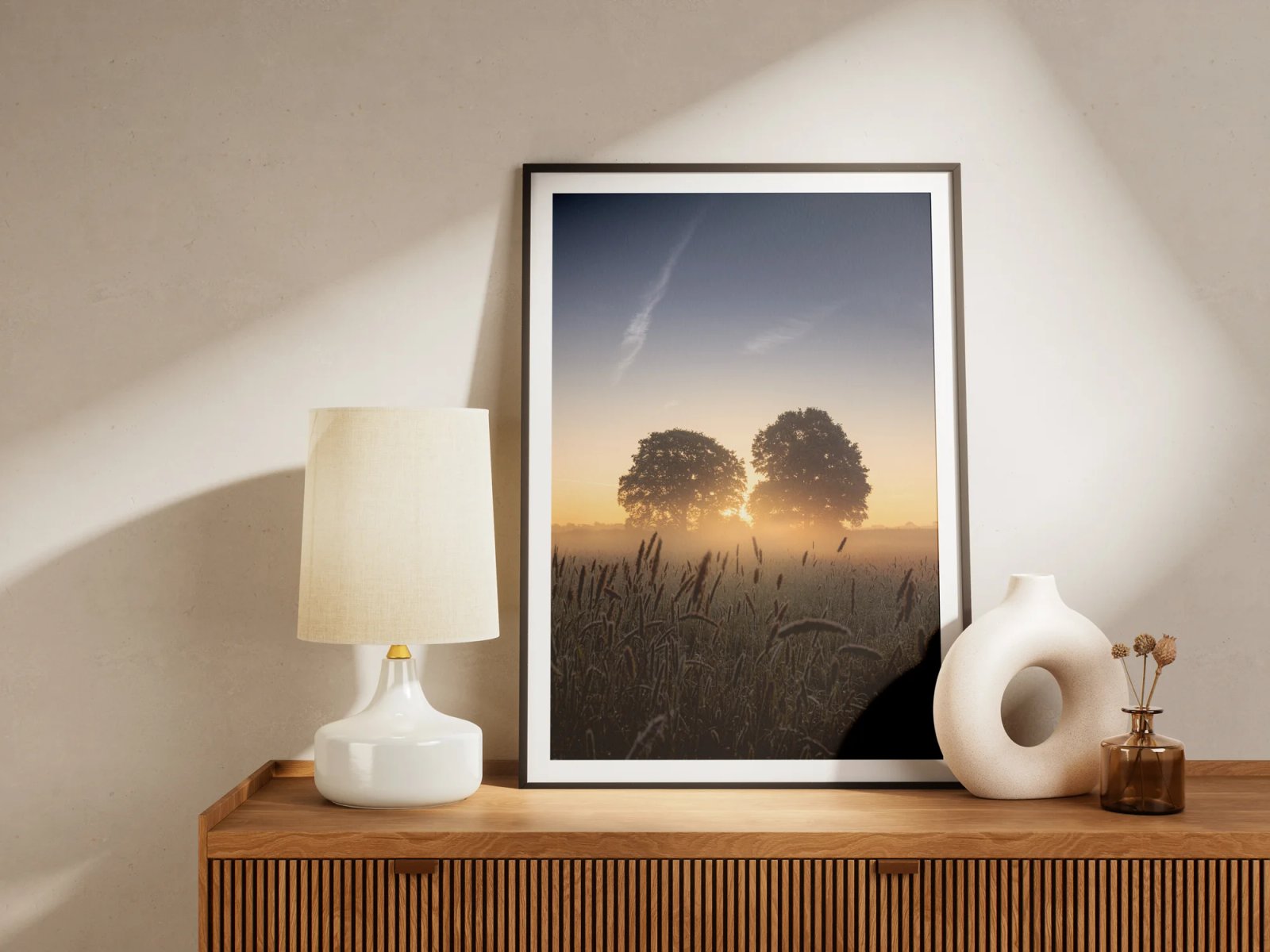 Der gerahmte Druck mit zwei Eichen im Morgenlicht über einem Feld steht auf einem Sideboard neben einer Lampe