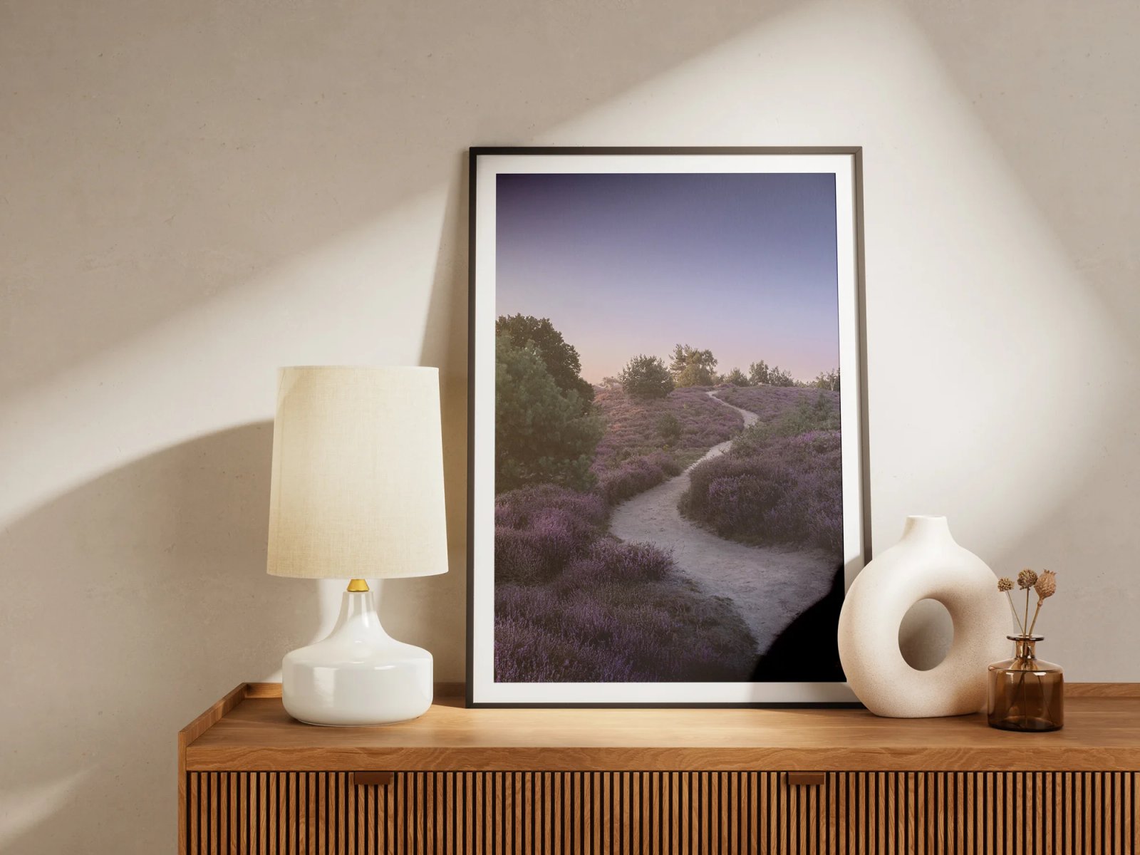 Der gerahmte Druck mit einem Sandweg durch blühende lila Heide in der Abenddämmerung steht auf einem Sideboard zwischen Lampe und Vase