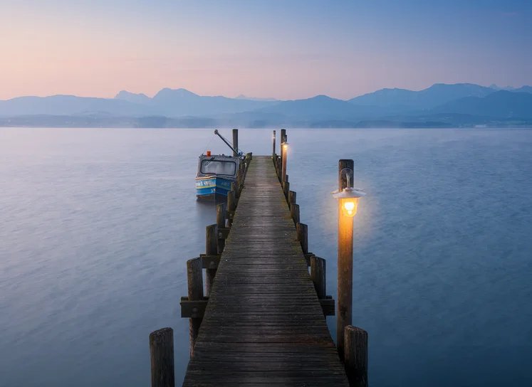 Ein Holzsteg mit Laterne führt in einen ruhigen See, dahinter Berge in der Morgendämmerung
