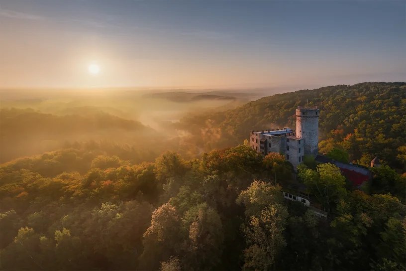 Eine Burg ragt aus einem herbstlichen Wald, dahinter geht über dem Nebel die Sonne auf