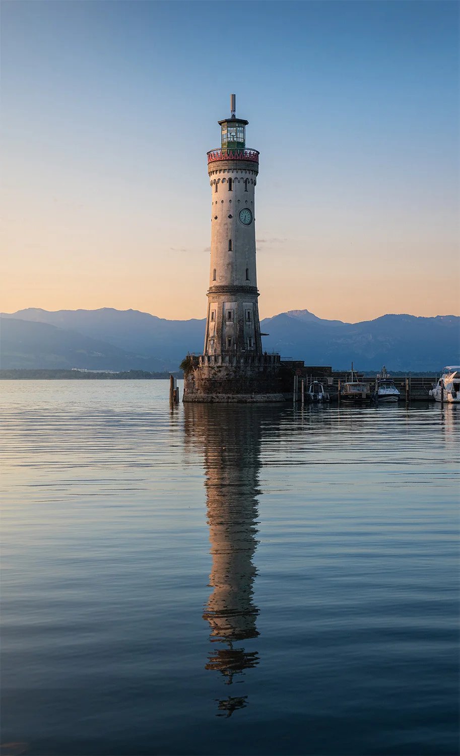Ein Leuchtturm spiegelt sich im ruhigen Wasser, am Horizont liegen Berge im Morgenlicht