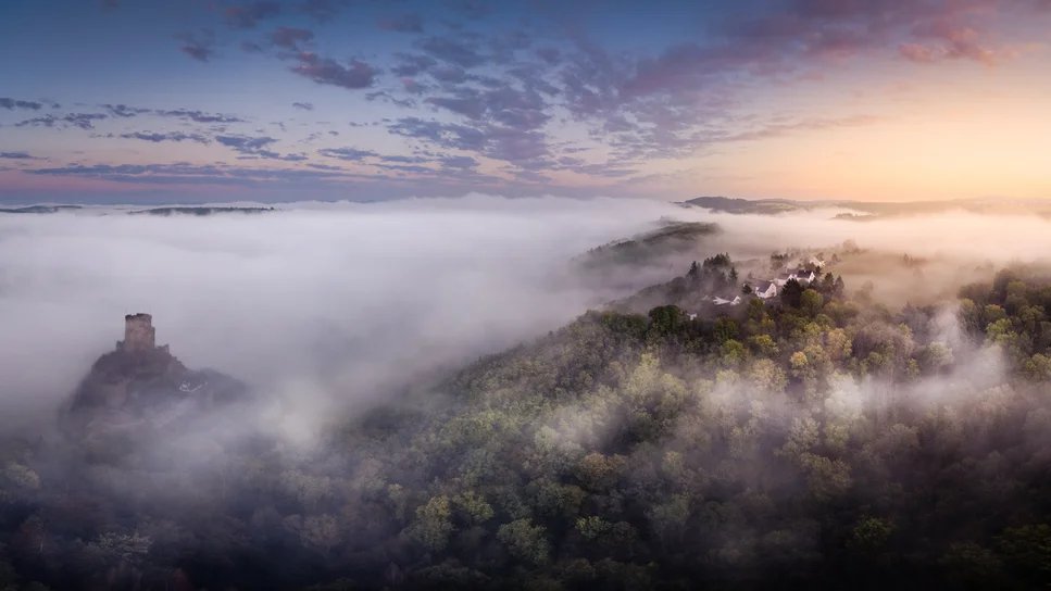 Burgruinen ragen bei Sonnenaufgang aus einem Nebelmeer über bewaldeten Hängen