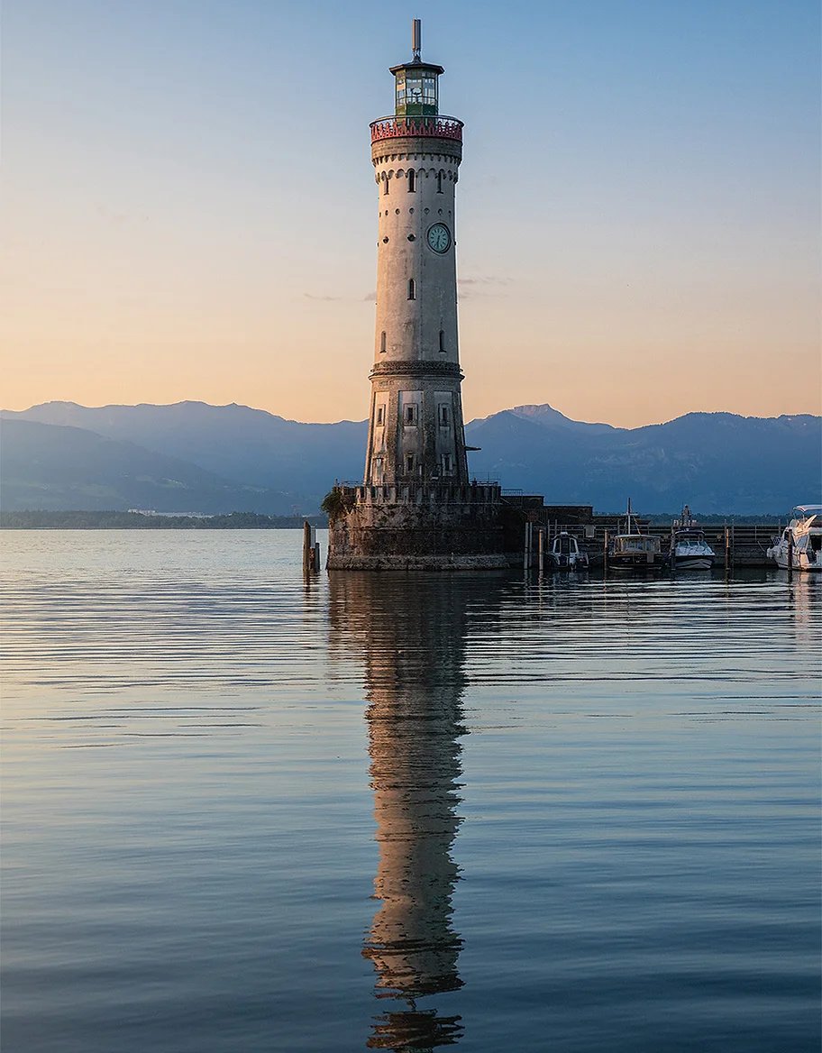 Ein Leuchtturm spiegelt sich im ruhigen Wasser, am Horizont liegen Berge im Morgenlicht