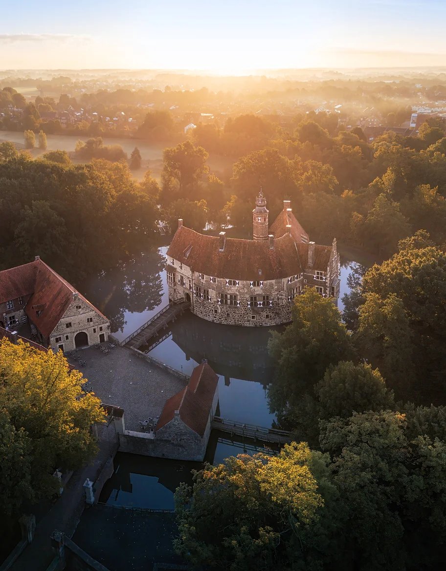 Luftaufnahme einer Wasserburg zwischen Bäumen, dahinter geht die Sonne auf
